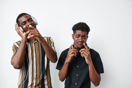 Two young black men with dark skin enjoying music together, one wearing headphones and striped shirt, the other in black shirt, expressing emotions and having fun on white background.の写真素材