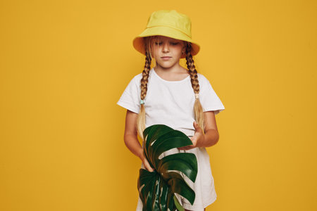 Young girl with braided hair wearing a yellow hat and white dress holding green tropical leaves on a yellow background.の写真素材