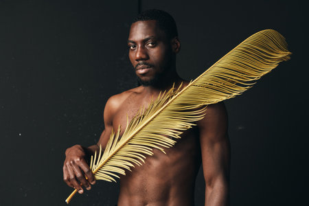 Black man with dark skin holding large golden palm leaf in creative visual designer style, artistic...