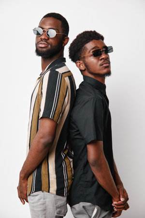 Two African American men with dark skin wearing sunglasses and casual shirts standing back to back against white background. Young adults with confident expressions and modern style.の写真素材