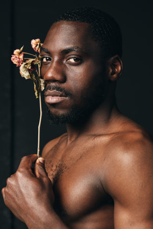 African man with dark skin holding dried flower close to face in creative conceptual visual style. Portrait, bare chest and serious expression on black background for artistic photography.の写真素材