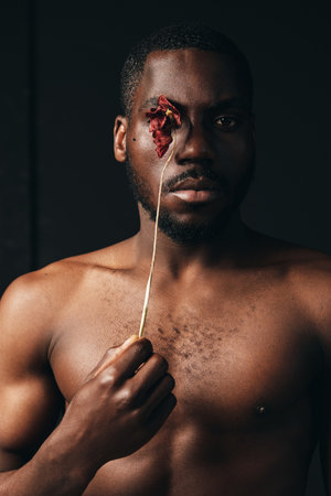 Creative conceptual portrait of a shirtless black man holding a dried flower over one eye in a dark studio. Artistic visual style with strong emotional expression and minimalistic background.の写真素材