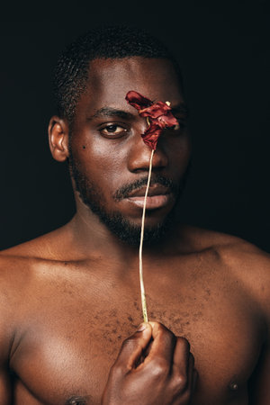 African man with dark skin holding dried flower in front of face in creative conceptual visual style. Portrait with bare chest and artistic expression on black background.の写真素材