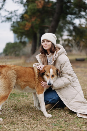 Woman kneels beside a dog in an open park, wearing a warm coat and beanie, offering gentle...