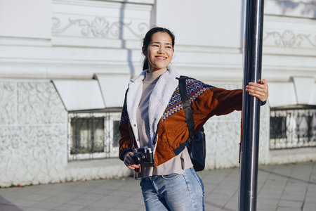 Woman with camera smiles beside a street pole, wearing a knit scarf, colorful jacket, and a...