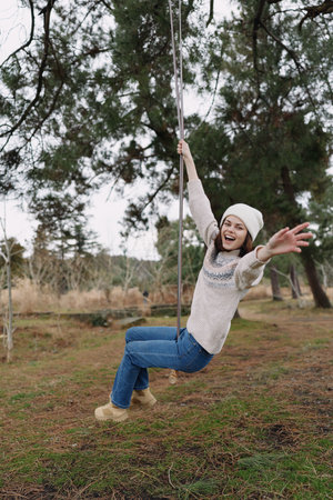 Young child on a swing in a park, outdoors, enjoying outdoor play, wearing a knit sweater and beanie, arms raised, smiling, carefree and full of fun, trees framing the scene.の写真素材