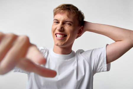 Joyful young man in a white T shirt smiles widely while pointing toward the camera, capturing a...