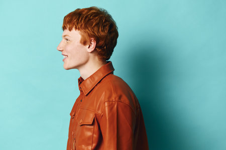 Profile of young man with red hair and freckles wearing brown leather jacket against blue...