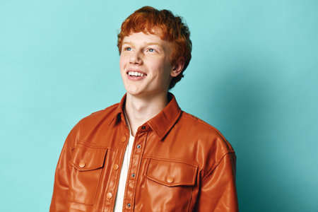 Young man with red hair wearing orange leather jacket smiling and looking away on blue background. Portrait of cheerful casual male with light skin and trendy outfit in studio.の写真素材