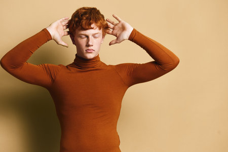 Young man with red hair in brown turtleneck poses with closed eyes and hands near head in minimal...