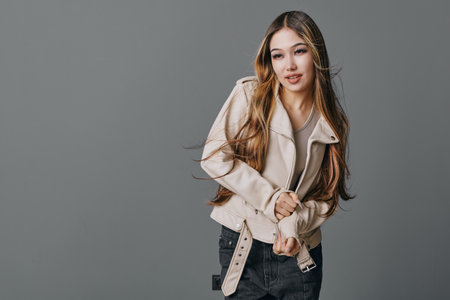 Young woman with long hair wearing a beige jacket and dark jeans posing on gray background. Female...