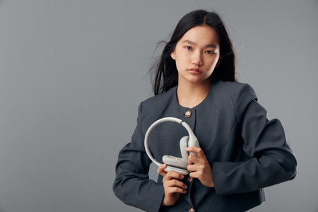 Young woman in gray jacket holding headphones in hand posing on gray background. Serious female...