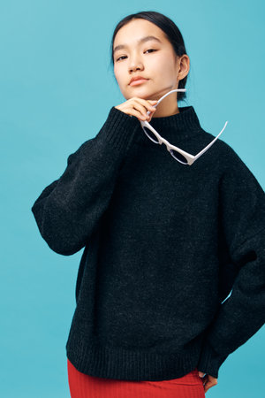 Young woman in black sweater holding white sunglasses posing confidently with hand near face against blue background. Fashion portrait of stylish casual outfit and natural makeup.の写真素材
