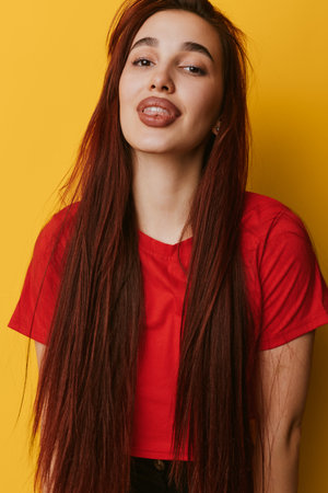 Young woman with long brown hair wearing red shirt posing with playful expression against yellow background. Casual style, confident female portrait with natural makeup and relaxed pose.の写真素材