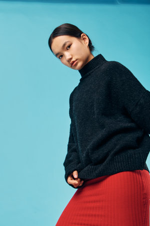 Young woman in a black sweater and red skirt posing against blue background, looking serious with...