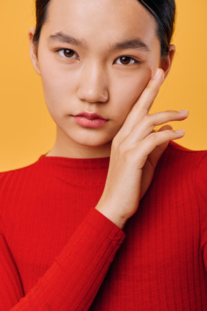 Young woman wearing red sweater posing with hand near face looking confident and calm on yellow background in studio portrait for fashion or beauty concept.の写真素材
