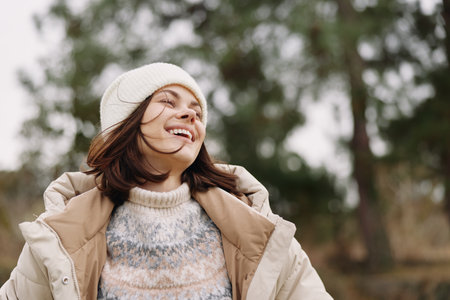 Woman outdoors in winter wind, smiling with a cozy beanie and puffer coat, enjoying fresh air among...