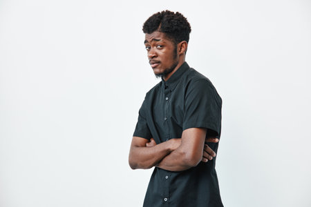 African American man with dark skin and curly hair wearing black shirt standing with arms crossed looking sideways on white background. Portrait, casual style and confident expression.の写真素材