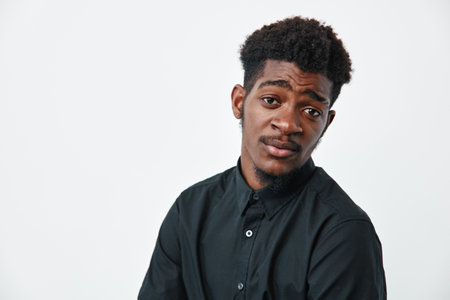 Young black man with dark skin and afro hairstyle wearing black shirt posing with thoughtful expression on white background. Male model with casual style and natural look.の写真素材