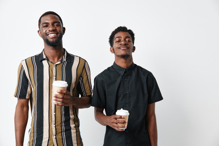 Two young African American men with dark skin holding coffee cups smiling and standing against white background in casual clothing for friendship and lifestyle.の写真素材