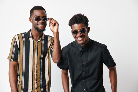 Two young black men with dark skin wearing sunglasses smiling and posing together in casual shirts...