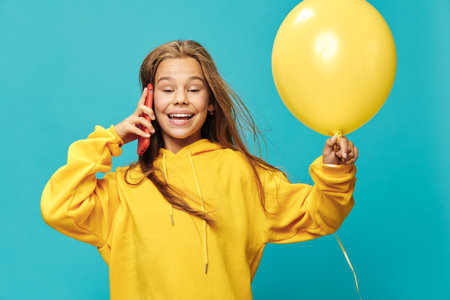 Happy girl in yellow hoodie holding balloon and talking on phone. Smiling child with closed eyes expressing joy and playfulness on blue background for fun and communication.の写真素材