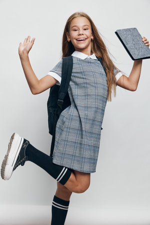 Girl student jumping and smiling with backpack and tablet in hand for education, learning and school concept. Child wearing uniform with happy expression and energetic pose on light background.の写真素材
