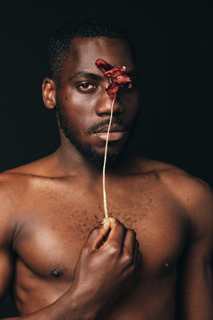 Creative conceptual portrait of African man with bare chest holding dried flower in front of face, dark background, artistic visual style, closeup, emotion and expression.の写真素材