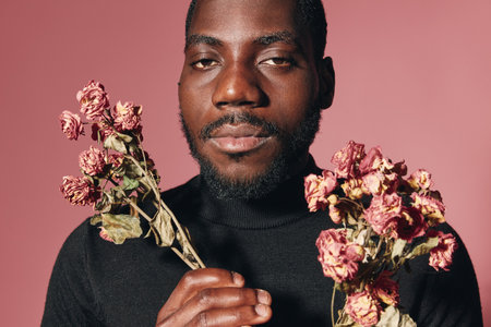 African man with beard holding dried pink flowers in both hands against pink background in creative conceptual visual style portrait with serious expression and black sweater.の写真素材