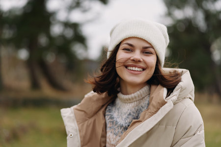 Woman outdoors in a winter coat and white beanie, smiling at the camera in a park setting,...