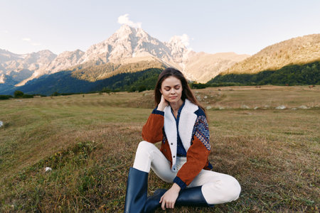 Woman portrait sitting in meadow with mountains in background, wearing shearling jacket and rubber boots, autumn outdoor scene, peaceful nature, relaxed young traveler.の写真素材