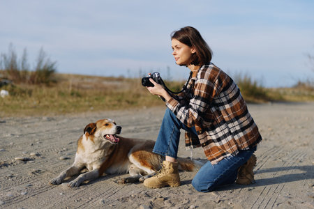 Woman photographer kneels on a sandy surface wearing a plaid jacket and boots capturing a joyful dog with a handheld camera during golden hour outdoor pet portrait and lifestyle momentの写真素材