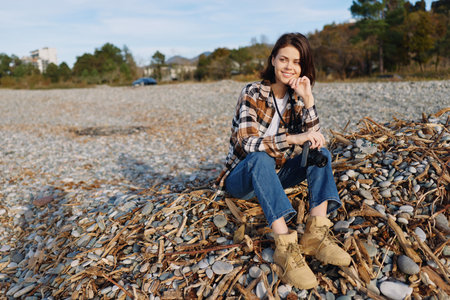 Woman on beach sits among rocks and pebbles in casual autumn attire, smiling softly under natural...