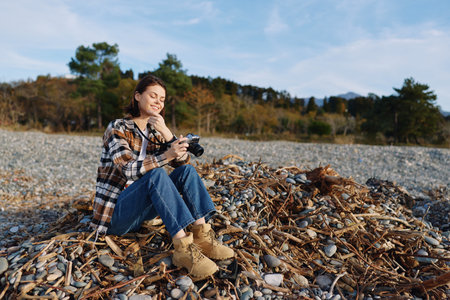 Photographer woman sitting on a rocky beach with a camera, wearing plaid shirt and boots, capturing a calm autumn shoreline with pebbles and distant trees under a clear sky.の写真素材