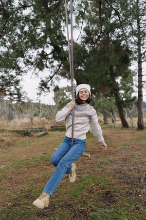 Girl on a swing in a park, enjoying outdoors. Cozy sweater and beanie, smiling as she swings from rope among trees, capturing a carefree childhood moment of joy and play.の写真素材