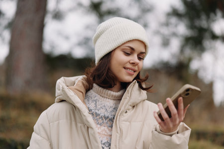 Woman in a winter coat and beanie checks her smartphone outdoors, smiling softly as she uses a...