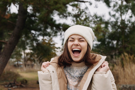 Woman in winter jacket outdoors, laughing with open mouth, wearing knit hat, a park scene around her, bright energy and candid happiness captured in a cool seasonal setting.の写真素材