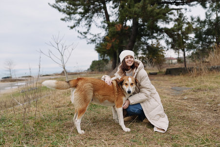 Woman in a warm coat hugging a friendly dog in an outdoor park with grass and a calm lake in the...