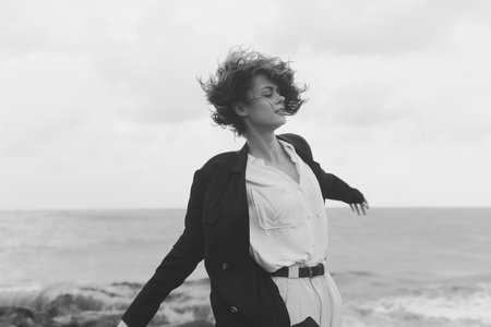 Monochrome image of a woman in a jacket and white shirt on a windy coast by the sea, hair blown,...