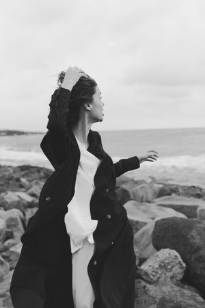 Wind swept figure on a rocky shore in a monochrome coat, catching sea breeze with dynamic movement,...