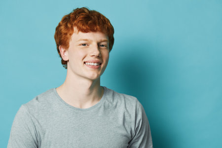 Young man with red hair smiling in casual gray shirt against blue background. Portrait of cheerful male with freckles and short hairstyle, relaxed and confident expression in studio.の写真素材