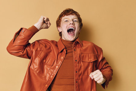 Young man with red hair wearing orange leather jacket and turtleneck celebrating with raised fist and open mouth on beige background.の写真素材