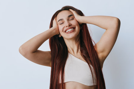 Young woman with long hair smiling with eyes closed and hands on head, wearing casual sleeveless top against plain background. Happy female portrait with relaxed joyful expression.の写真素材