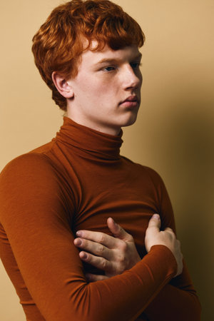 Portrait of young redhead man in brown turtleneck sweater with arms crossed on chest looking aside on beige background. Indoor studio shot with soft light on face and thoughtful expression.の写真素材