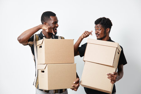Two african men with dark skin holding cardboard boxes and smiling while standing against white background for delivery and moving concept.の写真素材