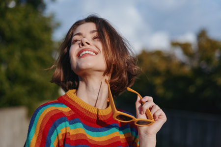Woman smile sunglasses rainbow sweater portrait outdoors, joyful young woman looking up and holding yellow sunglasses, candid fashion portrait with colorful knit and warm natural light.の写真素材