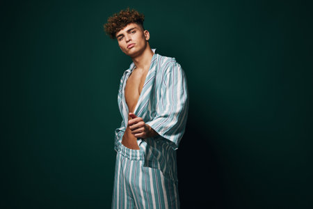 Young man with curly hair wearing striped pajamas posing confidently against dark green background. Casual style, relaxed mood, and modern fashion with open shirt and matching pants.の写真素材