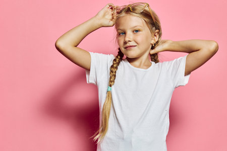 Blonde girl in a white t-shirt poses confidently with braided hair against a pink background. Studio portrait of a child expressing self-assurance and calmness.の写真素材