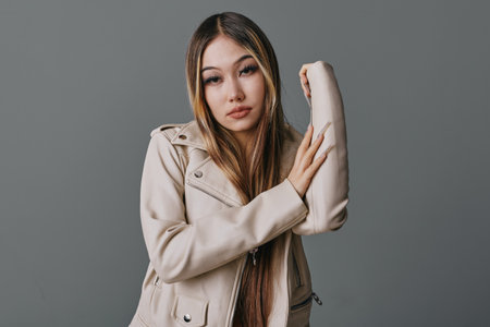 Young woman with long straight hair wearing beige jacket posing with raised arm against gray background, looking at camera with calm and confident expression in studio portrait.の写真素材