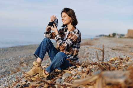 Woman sitting on a rocky beach with a camera, capturing a moment outdoors. Casual plaid shirt and...